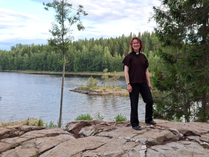 Hanne Keijonen by kotialbumi Nainen papinpaidassa seisoo kalliolla järvimaiseman edustalla. Taustalla näkyy metsää.