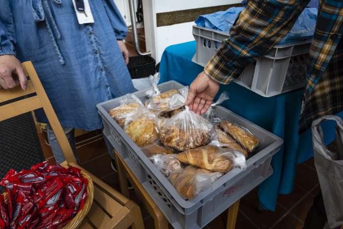 Leipiä harmaassa muovikorissa. Taustalla näkyy tummansiniseen mekkoon pukeutuneen henkilön torso ja etulalla käsi ottamassa yhtä leipäpussia.