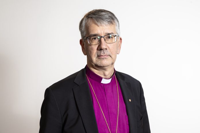 Harmaahapsinen mies violetissa piispan pantapaidassa. Valkoinen tausta.