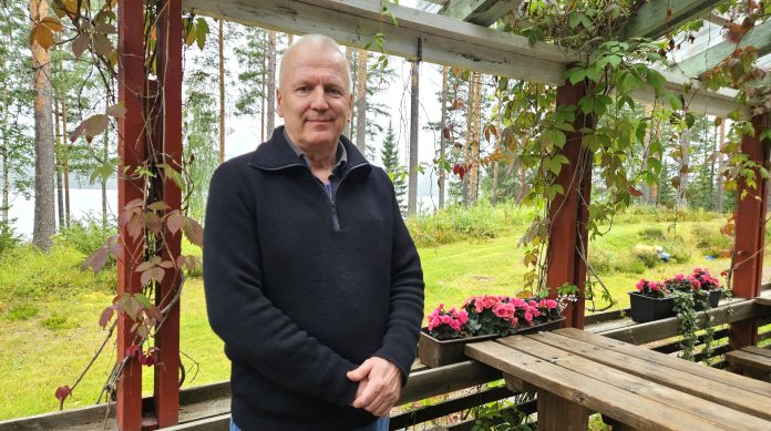 Vesa Hirvonen by Emilia Karhu Kuvassa puolilähikuva miehestä jolla on tumma villatakki. Taustalla näkyy puita ja terassin kukkalaatikko.