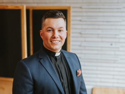 Hymyilevä henkilö tummansinisessä puvussa ja papinkauluksessa seisoo vaaleassa puuverhoillussa sisätilassa. Puvun rintataskussa on värikäs taskuliina.