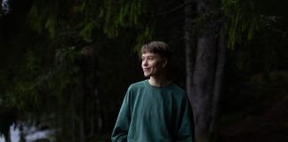 Lyhythiuksinen henkilö seisoo vihreässä collegepaidassa hämärässä metsässä kädet taskuissa ja katsoo sivulle hymyillen. Taustalla näkyy havupuita ja rantaa.