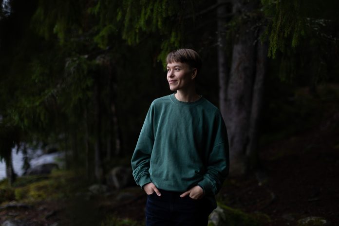 Pilvi Hyväri Lyhythiuksinen henkilö seisoo vihreässä collegepaidassa hämärässä metsässä kädet taskuissa ja katsoo sivulle hymyillen. Taustalla näkyy havupuita ja rantaa.