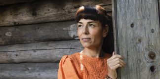 Kuvassa nainen katsoo puolilähikuvassa tummahiuksinen ja oranssimekkoinen vasemmalle. Taustalla näkyy kelopuuseinä.