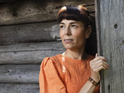 Kuvassa nainen katsoo puolilähikuvassa tummahiuksinen ja oranssimekkoinen vasemmalle. Taustalla näkyy kelopuuseinä.