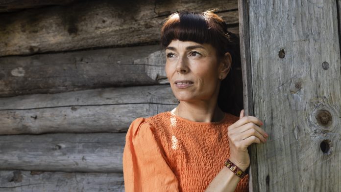 Kral-Leszczynski Alexandra-9394 by Jukka G Kuvassa nainen katsoo puolilähikuvassa tummahiuksinen ja oranssimekkoinen vasemmalle. Taustalla näkyy kelopuuseinä.