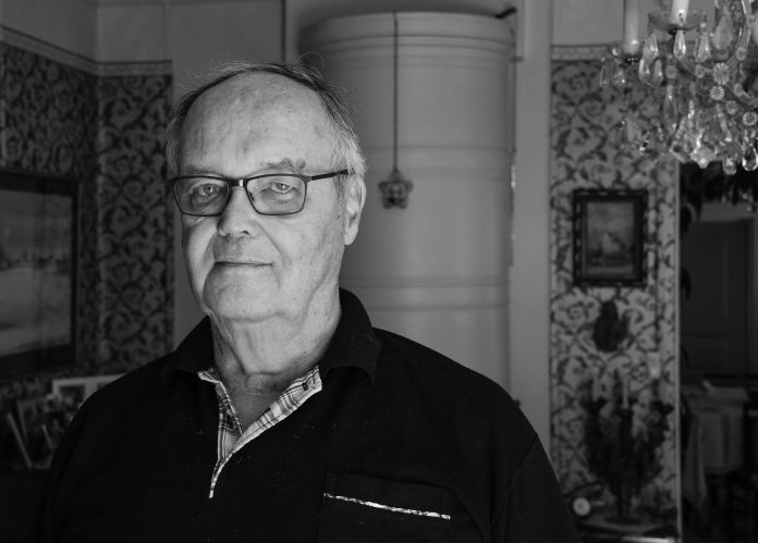 ALT-teksti: Silmälasipäinen mies katsoo kameraan mustavalkoisessa muotokuvassa. Taustalla näkyy koristeellisesti tapetoitu huone, tauluja ja kristallikruunu.