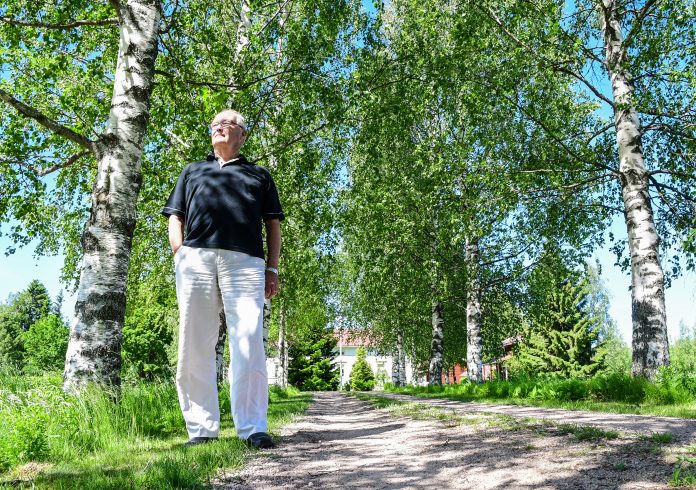 Iäkäs mies seisoo koivujen keskellä aurinkoisella pihalla. Hänellä on musta paita ja valkoiset housut, ja taustalla näkyy vehreää kasvillisuutta, hiekkatie ja rakennuksia.