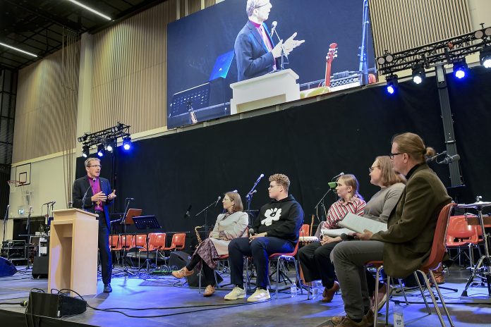 Piispa puhuu puhujakorokkeella sisätilaisuuden lavalla viidelle tuoleilla istuvalle henkilölle. Taustalla näkyy mikrofoneja, soittimia ja tyhjiä tuoleja, ja suuri näyttö välittää lähikuvaa piispasta.