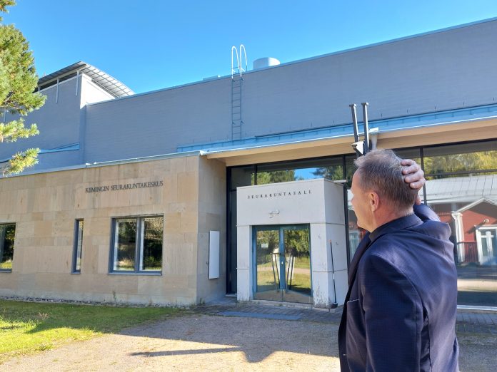 Seurakuntakeskus by Tapio Mainio Mies seisoo aurinkoisella pihalla selin kameraan ja katsoo vaaleaa rakennusta, jonka seinässä lukee ”Kiimingin seurakuntakeskus”. Sisäänkäynnin yläpuolella on teksti ”Seurakuntasali”. Mies pitää toista kättään takaraivollaan.