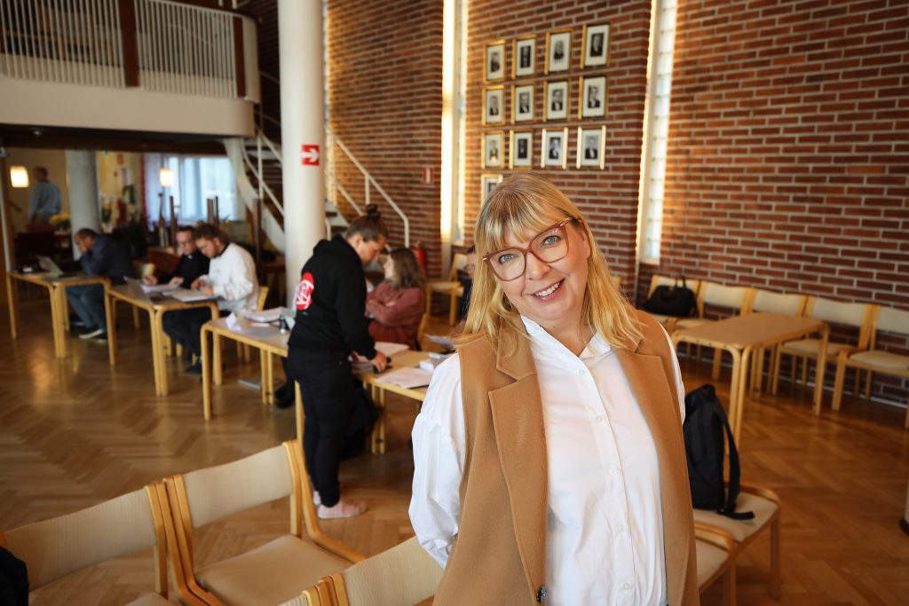 naine seisoo seurakuntassalin keskellä ja katsoo hymyillen kameraan