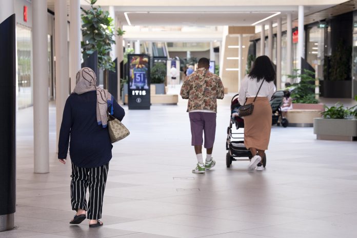 Kuluttajia Itäkeskuksessa Ihmisiä kävelee valoisan ja avaran kauppakeskuksen käytävällä. Etualalla huiviin pukeutunut henkilö kulkee poispäin kamerasta, ja taaempana kaksi muuta henkilöä kävelee samaan suuntaan, toinen lastenvaunuja työntäen. Käytävää reunustavat viherkasvit ja liiketilat.