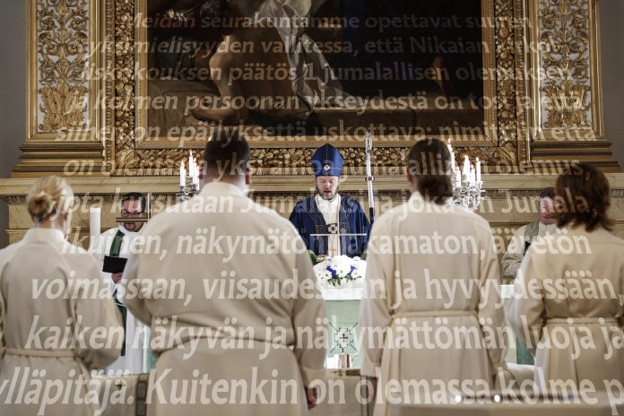 Timo Laanisen pappisvihkimys Kirkossa toimitettava jumalanpalvelus. Alttarin edessä seisoo vaaleisiin asuihin pukeutuneita henkilöitä, ja keskellä pappi lukee alttarilta. Taustalla on suuri kehystetty maalaus ja kynttilöitä. Kuvan päällä kulkee suurikokoista, osittain läpikuultavaa suomenkielistä tekstiä.