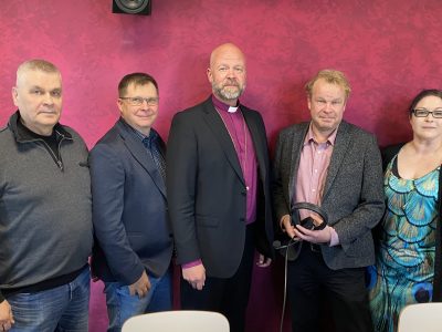 Viisi henkilöä seisoo punaise taustan edelssä. Keskimmäisellä piispan violetti paita.