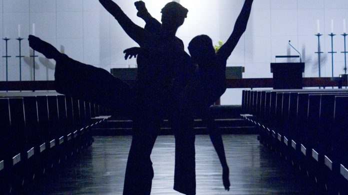 Xaris Dance -tanssiryhmä Kolmen tanssijan tummat siluetit kirkkosalissa. Tanssijat muodostavat yhdessä dynaamisen asennon: kaksi ojentaa jalkansa sivuille ja yksi kohottaa kätensä korkealle. Taustalla näkyy alttari ja riveittäin tyhjiä penkkejä.