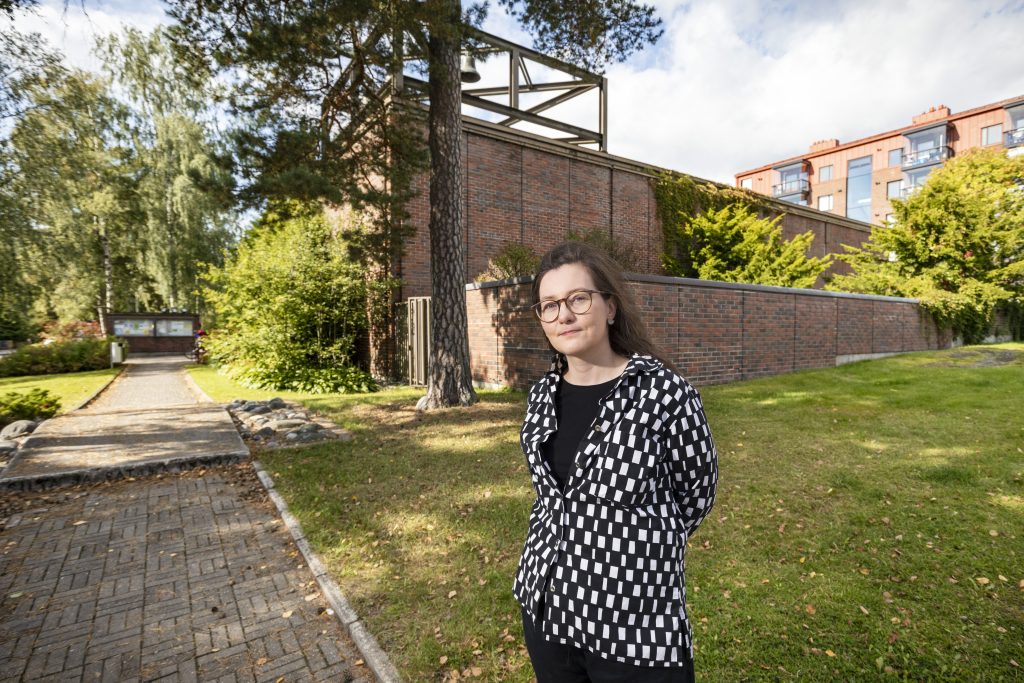 Silmälasipäinen nainen seisoo nurmikolla mustavalkoisessa kuviollisessa takissa. Taustalla on punatiilinen rakennus, korkea mänty, muita puita ja kävelytie.