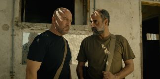 "Two serious men with shoulder bags stand in front of a weathered, sunlit building, partially shaded by window light inside a worn, possibly abandoned environment."