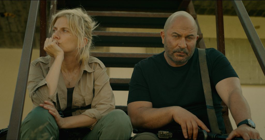 "Two serious people sit on outdoor metal stairs in casual, tactical clothing, appearing tired or contemplative, with sunlight casting shadows behind them against a plain wall in a rugged environment."