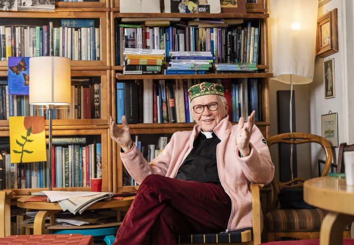 Hynninen Veli-Matti-0346 by JG Iloinen mies istuu kirjahyllyn edustalla värikäs hattu päässään ja kädet levällään. Päällä musta papin paita ja vaaleanpunainen avonainen kauluspaita.