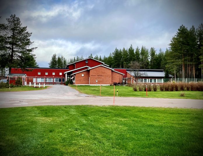 Kestilän vankila by Rikosseuraamuslaitos Punainen, osittain tiiliverhoiltu rakennus, jonka julkisivussa lukee ”Kestilän avovankila”. Rakennuksen edessä on vihreä nurmialue ja hiekkatie. Taustalla kohoaa mäntymetsä, ja taivas on pilvinen.