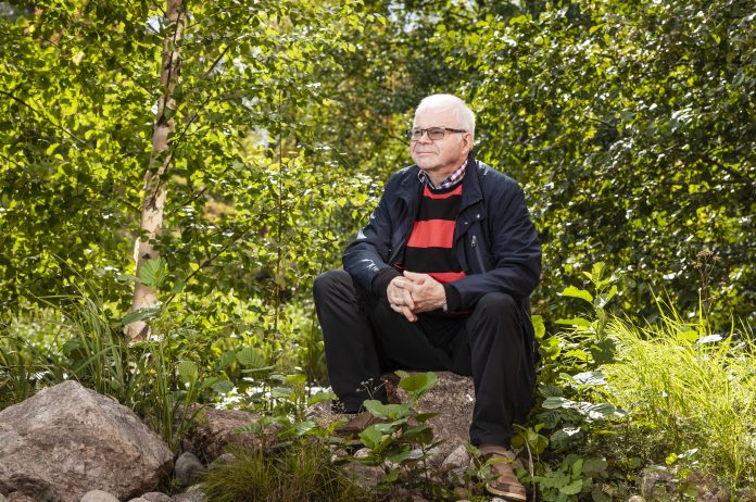 Kettunen Paavo-0691 by JG Valkohiuksinen, silmälasipäinen mies istuu kivellä vehreässä metsämaisemassa. Hänellä on tumma takki ja punamustaraidallinen paita. Mies pitää käsiään ristissä polvien välissä ja katsoo hieman sivulle. Ympärillä kasvaa koivuja, pensaita ja ruohoa.