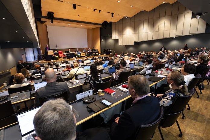 Näkymä istuntosaliin takaa päin kirkolliskokouksessa. Riveissä ihmisiä, joiden edessä mikrofonit, tietokoneita ja muistiinpanovälineitä.