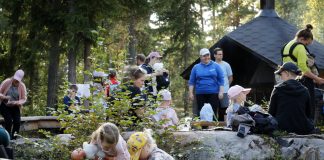 Metsäisessä ympäristössä on koolla lapsia ja aikuisia. Etualalla kaksi pientä lasta tutkii maaston kasvillisuutta, ja toisella on nukke kainalossaan. Taustalla muita osallistujia istuu ja seisoo nuotiokatoksen ympärillä.