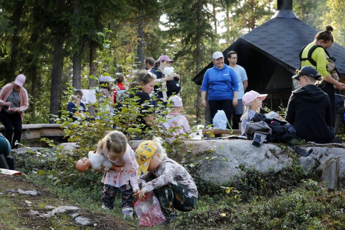 Metsäisessä ympäristössä on koolla lapsia ja aikuisia. Etualalla kaksi pientä lasta tutkii maaston kasvillisuutta, ja toisella on nukke kainalossaan. Taustalla muita osallistujia istuu ja seisoo nuotiokatoksen ympärillä.