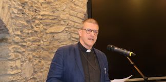 Siniseen pikkutakkiin ja mustaan papinpaitaan pukeutunut miespastori puhuu mikrofoniin luentotilassa.