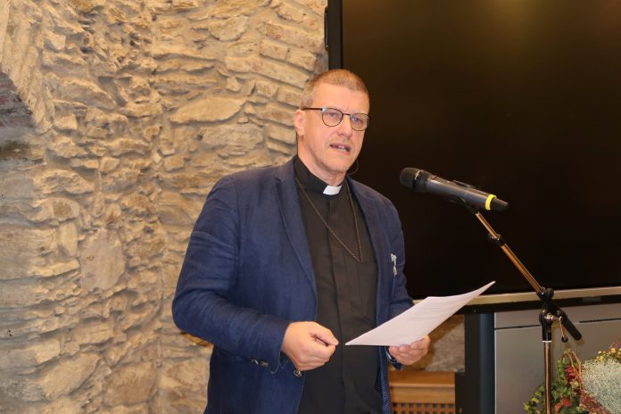 Siniseen pikkutakkiin ja mustaan papinpaitaan pukeutunut miespastori puhuu mikrofoniin luentotilassa.