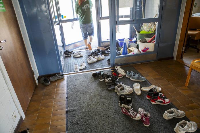 Saarijarvi_2 Eteistilassa tiililattia ja harmaa matto, jonka päällä sikin sokin nuorten kenkiä. Taustalla näkyy tuulikaappi ja lasiovi, josta joku on astumassa juuri ulos farkkushortseissa ja vihreässä hupparissa.