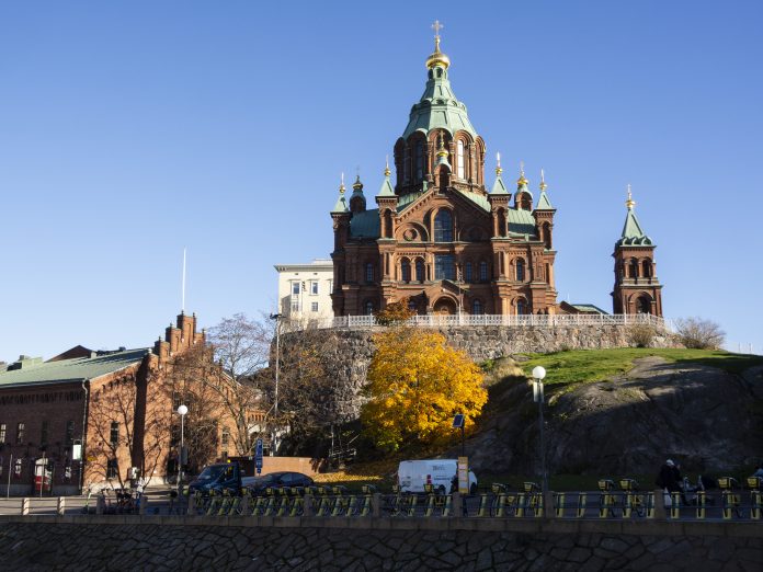 Uspenskin katedraali by JG-202574 Helsingin Uspenskin katedraali kohoaa kallioisella kukkulalla kirkkaansinistä taivasta vasten. Punatiilistä ortodoksikirkkoa koristavat vihreät katot ja kullatut kupolit risteineen. Etualalla näkyy keltaiseksi värjäytyneitä syyspuita, katua ja pysäköityjä ajoneuvoja.