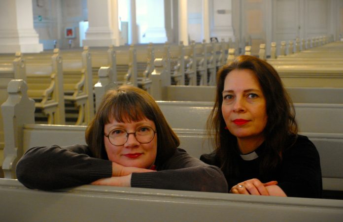Kaksi naista istuu vierekkäin vaalean kirkonpenkin takana ja katsoo kameraan. Vasemmalla silmälasipäinen, otsatukkainen nainen nojaa käsivarsiinsa. Oikealla tummahiuksisella naisella on musta papin paita ja valkoinen liperi. Taustalla näkyy pitkiä rivejä vaaleita, tyhjiä kirkonpenkkejä.