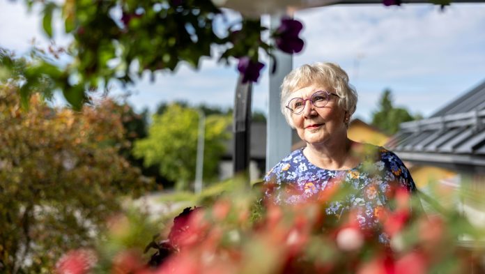 Kuvassa harmaahiuksinen silmälasipäinen nainen katsoo vasemmalle. Taustalla näkyy puita ja vettä.