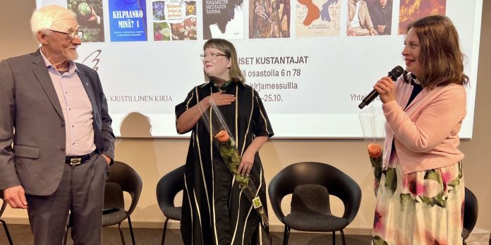 voittaja by Kaj Aalto Mies ja kaksi naista seisovat kirkkaan näytön edessä. Taustalla mustia tuoleja, molemmilla naisilla ruusut ja toisella naisista mikrofoni kädessä.
